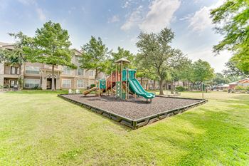 A playground with a green slide in the middle of a grassy area.